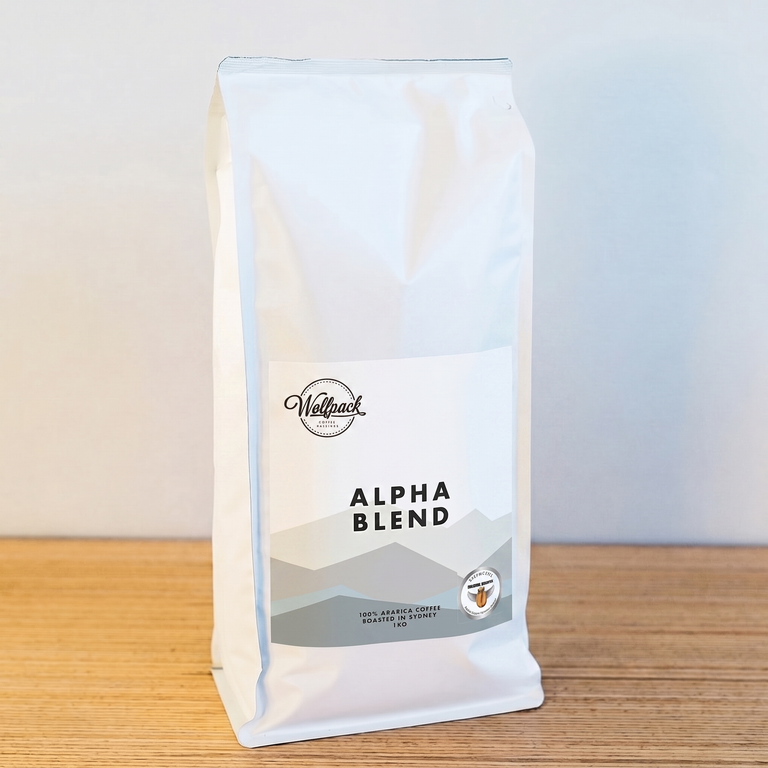 A white bag of Wellpack Alpha Blend coffee stands on a light wood surface against a plain light gray wall.